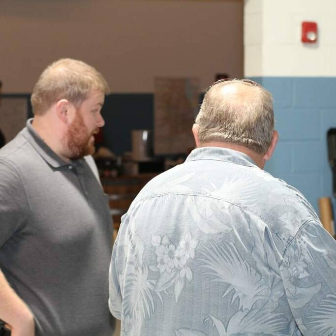 Two men are conversing indoors. The man on the left, wearing a gray polo shirt and sporting a beard, engages with the man on the right, who wears a light blue floral shirt. The room has blue and white walls with various equipment visible in the background—perhaps discussing Oktoberfest plans in September or an upcoming IACAC event.