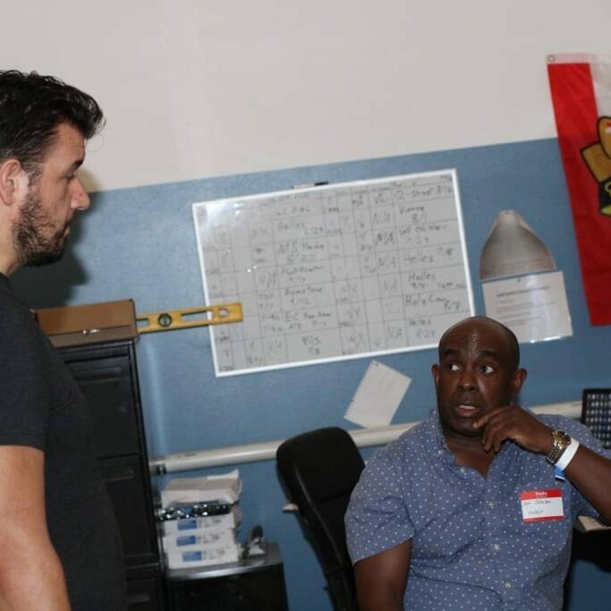 Two men are in a room. One is sitting at a desk, and the other is standing, engaged in conversation. A whiteboard covered with notes is in the background, next to a colorful flag with an emblem from the 2018 IACAC conference. The room has various office items and equipment.