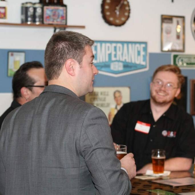 A group of men stands in a casual bar area, conversing. One man, in a gray suit, holds a drink and is facing away, while another man in a black shirt and glasses smiles at him. The background features various signs and posters celebrating Oktoberfest and a clock on the wall hinting at September’s arrival.