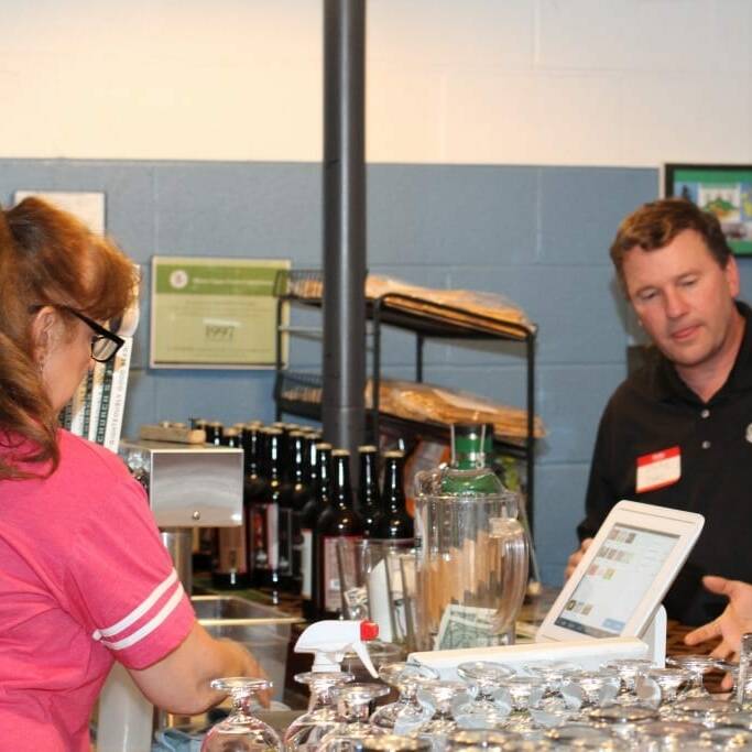 A woman in a pink shirt stands behind a counter with several glasses, facing a cash register. Two people, a man and a woman, are on the other side of the counter, engaged in conversation about Oktoberfest 2018. Shelves with bottles and a jar with tips for IACAC are visible in the background.