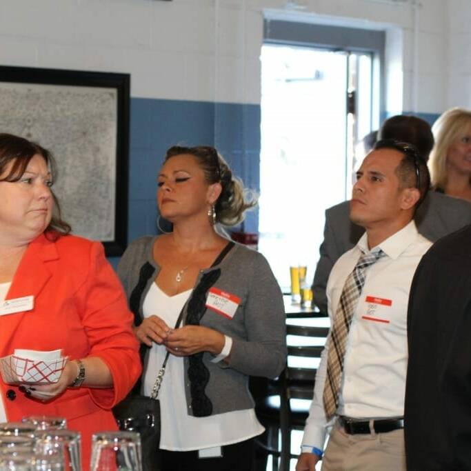 A group of people are gathered indoors, engaging in conversation and activities during the 2018 IACAC event. One woman is wearing a red blazer, and several people have name tags on. The background shows a large window and framed artwork on the wall. Glassware and tables are visible in the scene.