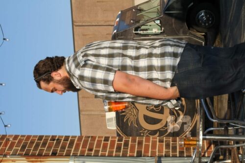 A person with a beard and long hair, wearing a plaid shirt and jeans, stands outside next to a brick wall during the 2018 IACAC. Their hands are in their pockets, and their head is slightly tilted forward. In the background, there's a van and a sign with a "D.