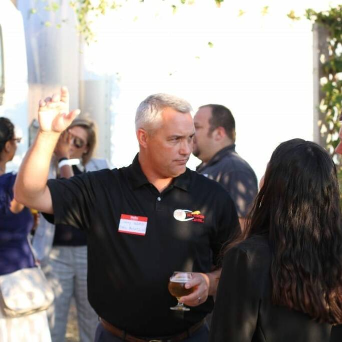 A man with short gray hair in a black polo shirt gestures with his hand while holding a drink and speaking to two individuals at an outdoor IACAC event. Several other people are visible in the background near a truck. The man wears a name tag, suggesting it's likely an early Oktoberfest celebration in September.