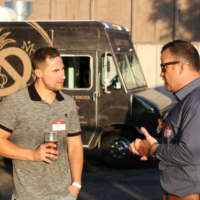 Two men stand outdoors near a food truck with a pretzel logo, having a conversation. One man, holding a glass, wears a grey polo shirt, while the other, gesturing with one hand, wears glasses and a blue button-up shirt. The scene feels festive as if captured during an Oktoberfest celebration in September.