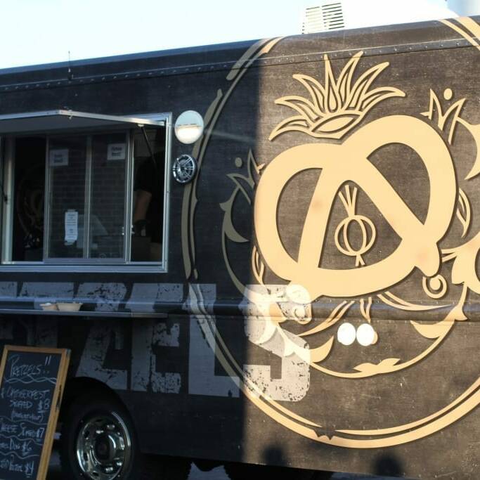 A black food truck adorned with a large golden pretzel logo and the slogan "EAT MORE PRETZELS" on the side. A menu board is visible near the serving window, listing various food items perfect for September's Oktoberfest. Sunlight casts shadows on the truck, highlighting the logo and text.