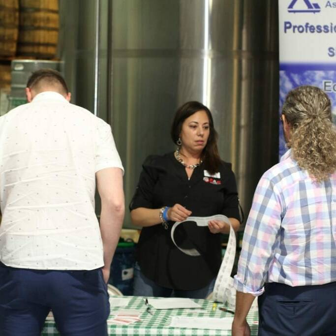 Three people are gathered at a table covered in a green checkered tablecloth at an indoor event. A woman behind the table is holding a paper and appears to be in conversation with two men who are facing away from the camera. A banner for 2018 IACAC's September Oktoberfest is in the background.