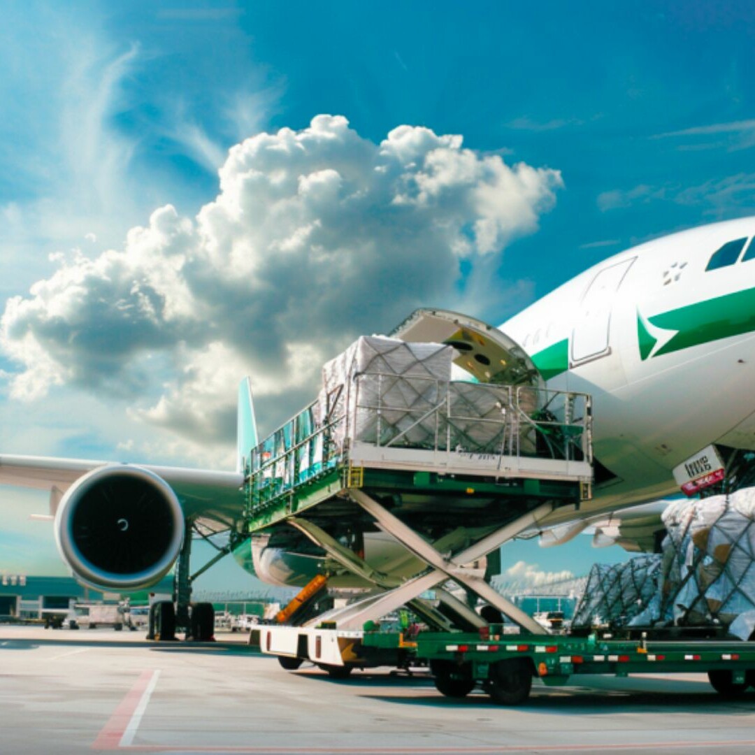 A cargo plane is being loaded with large bundles of goods at the airport, highlighting the efficiency of air cargo operations. The bundles, secured with nets, are transferred from a green and white loading vehicle. The sky is bright with scattered clouds in the background, suggesting a greener horizon ahead.