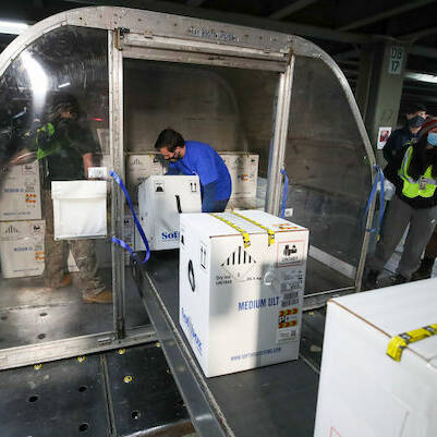 In December 2020, airport workers carefully load large white boxes labeled "Medium Upright" with temperature control symbols onto a plane's cargo hold. Wearing safety vests, masks, and gloves, they demonstrate a clear focus on safety and precaution.