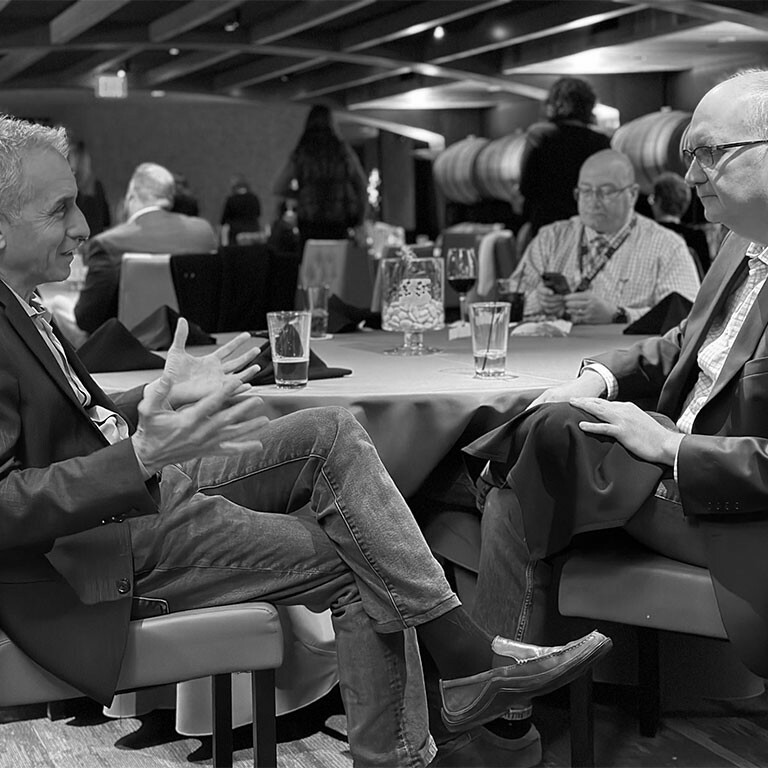 Two men in suits are engaged in a lively conversation at a round table, seemingly making contact over an important topic. They both have glasses of water in front of them. Other diners and tables are visible in the background, adding to the scene’s dynamism. The image is in black and white.