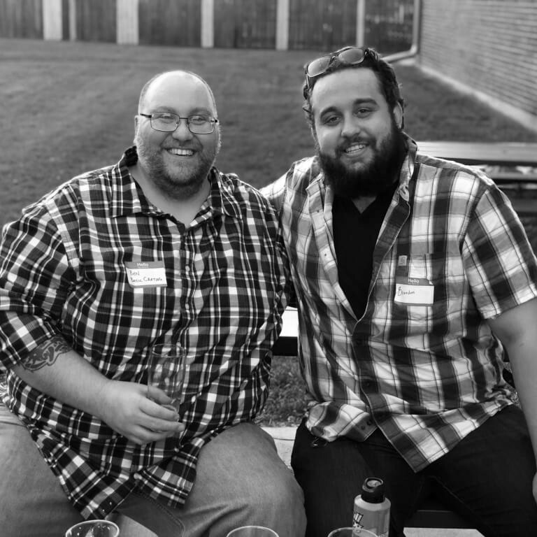 Two men sitting on a bench outdoors, both smiling and making eye contact with the camera. They are wearing checkered shirts and name tags, and one is holding a drink. There is grass and a brick wall in the background along with some picnic tables. The image is in black and white.