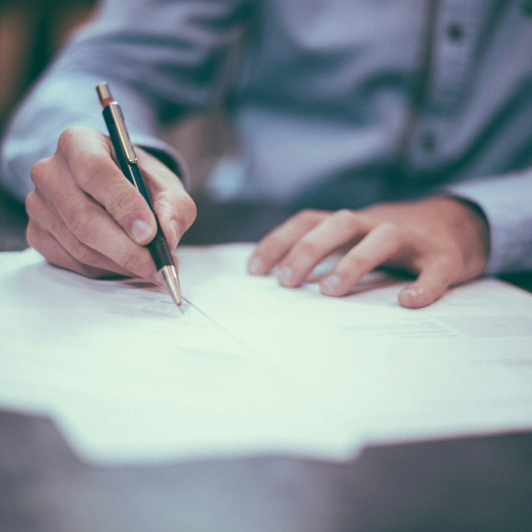 A person in a blue shirt writes on a piece of paper with a pen. Several sheets of paper, including the President's Letter from April 2020, are spread out on the table. The focus is on the hands and the pen, while the rest of the scene is slightly blurred.