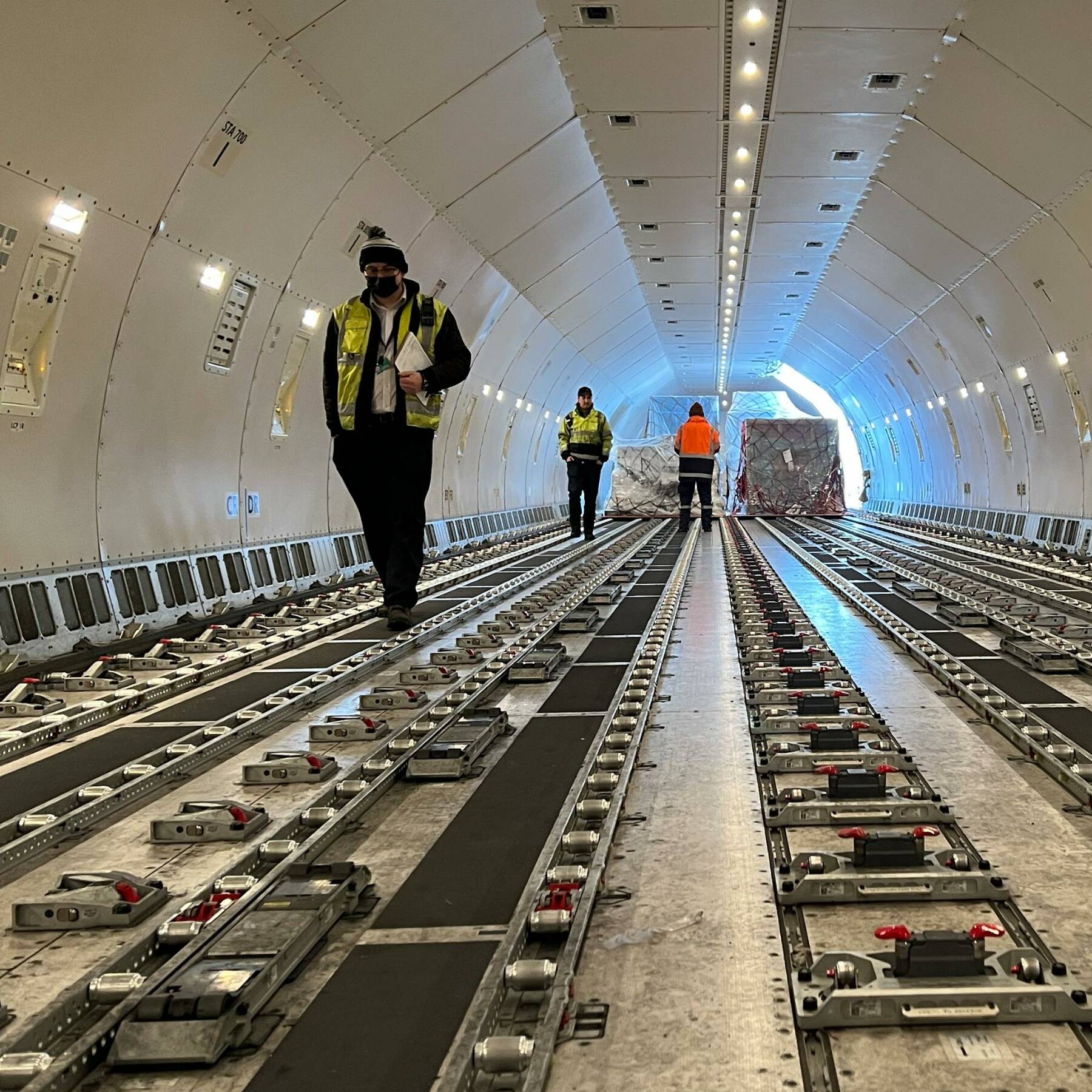 Workers in high-visibility clothing walk inside the empty cargo hold of an aircraft. The interior is lined with roller tracks for moving cargo, and the distant plane entrance is illuminated by natural light.