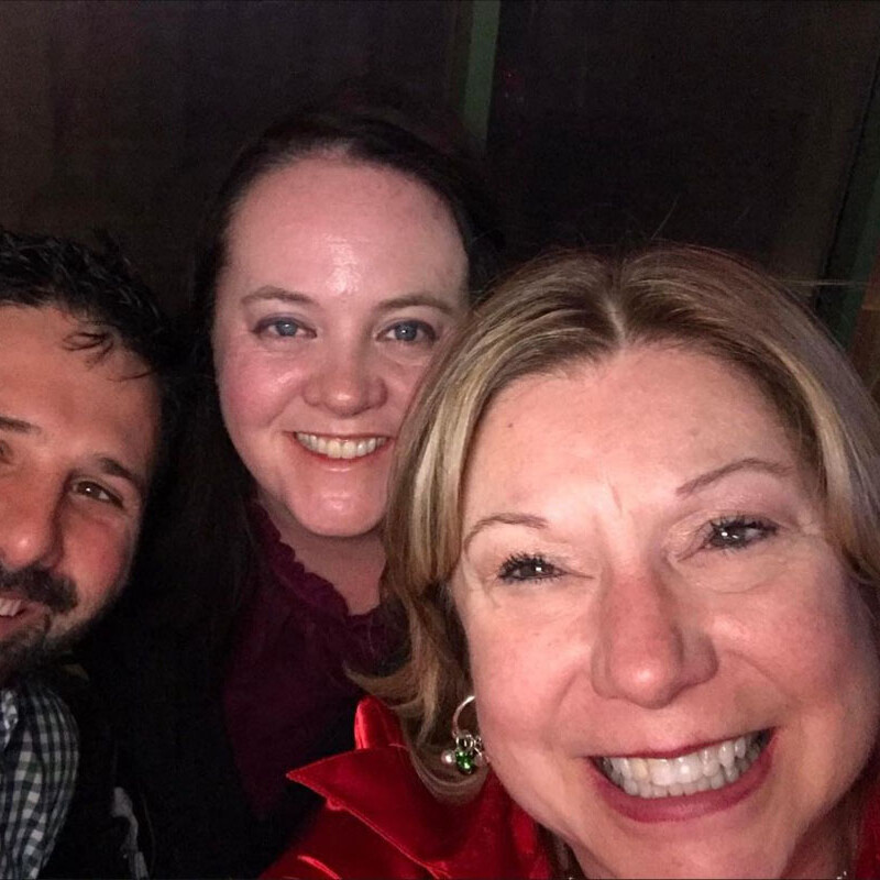 Three people smiling at the camera. The person on the left has dark hair and a beard, the person in the middle has dark hair, and the person on the right has light hair and is wearing a red garment with green earrings. They appear to be at a 2023 holiday party indoors in a well-lit area.