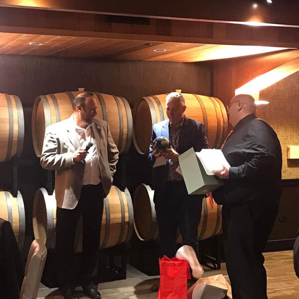 Three men are engaged in conversation in front of a wall stacked with wine barrels. It’s a lively 2023 holiday party. The man on the left wears a light blazer. The man in the center, holding a bottle, is dressed in a blue blazer. The man on the right, with a box, sports a dark suit and red tie.
