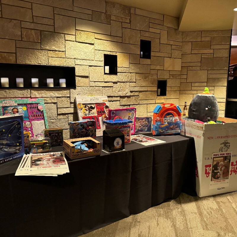 A display table covered with various boxed toys and games, including action figures, playsets, and dolls, is set against a stone wall backdrop. To the right of the table stands a large donation box labeled "Toys for Tots," filled with more holiday toys to spread cheer in 2023.