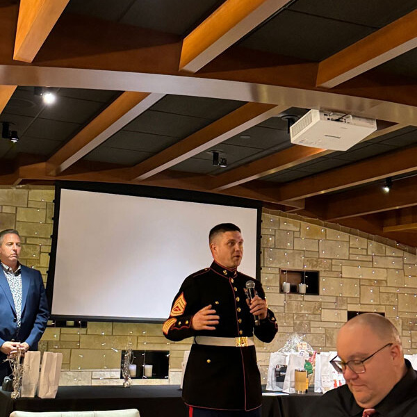 A man in formal military uniform speaks at a podium in a banquet hall, with another man standing to the left. A slide screen and various items are behind them. An audience member in a suit and glasses is seated in the foreground, capturing the spirit of the 2023 holiday party.