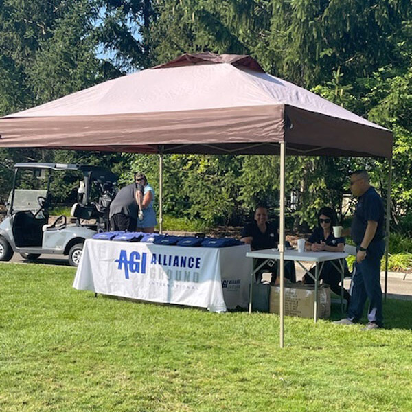 A brown tent with a table underneath displaying a white tablecloth with "AGI Alliance 2024" written on it. Three people are sitting or standing near the table. A golf cart and lush green trees are in the background, suggesting an outdoor golf outing.