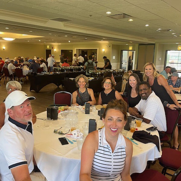 A group of people smiling and posing for a photo around a circular table in a banquet hall, possibly celebrating after the 2024 Golf Outing. The table has white cloths and some place settings. Other people are seen in the background engaged in conversation, adding to the warm and lively atmosphere.