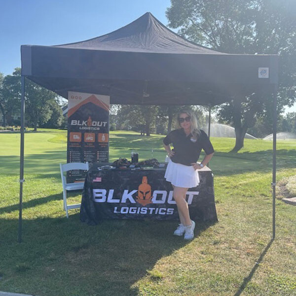 A woman stands under a black canopy tent displaying a table covered with a black tablecloth that reads "BLK OUT LOGISTICS." Behind her, there's a green grassy area with trees and a water feature under the bright, clear sky, perfect for an outdoor outing or a 2024 golf event.