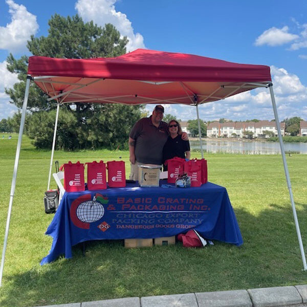 Two people stand under a canopy tent at an outdoor event. The table in front of them, covered with a blue tablecloth reading "Basic Crating Packaging Inc.," is filled with red bags, a clear box, and brochures. The background reveals a grassy area with a pond and houses, evoking the feel of a serene golf outing for 2024.