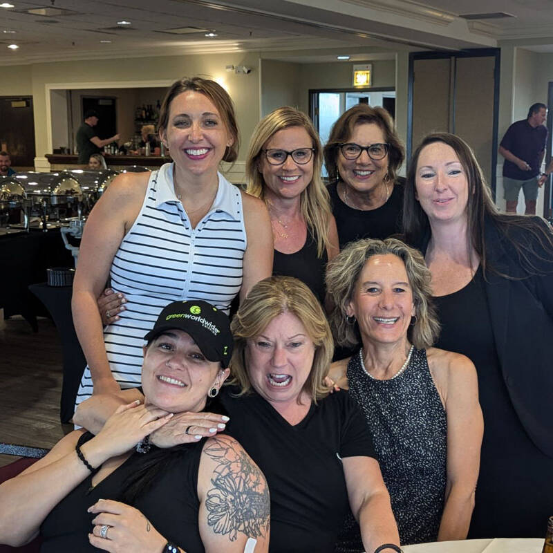 A group of seven people, mostly women, posing and smiling for a photo at a restaurant after a delightful golf outing. The group appears happy and relaxed, with some wearing informal yet stylish attire. Tables and buffet dishes are visible in the background, highlighting the celebratory ambiance.