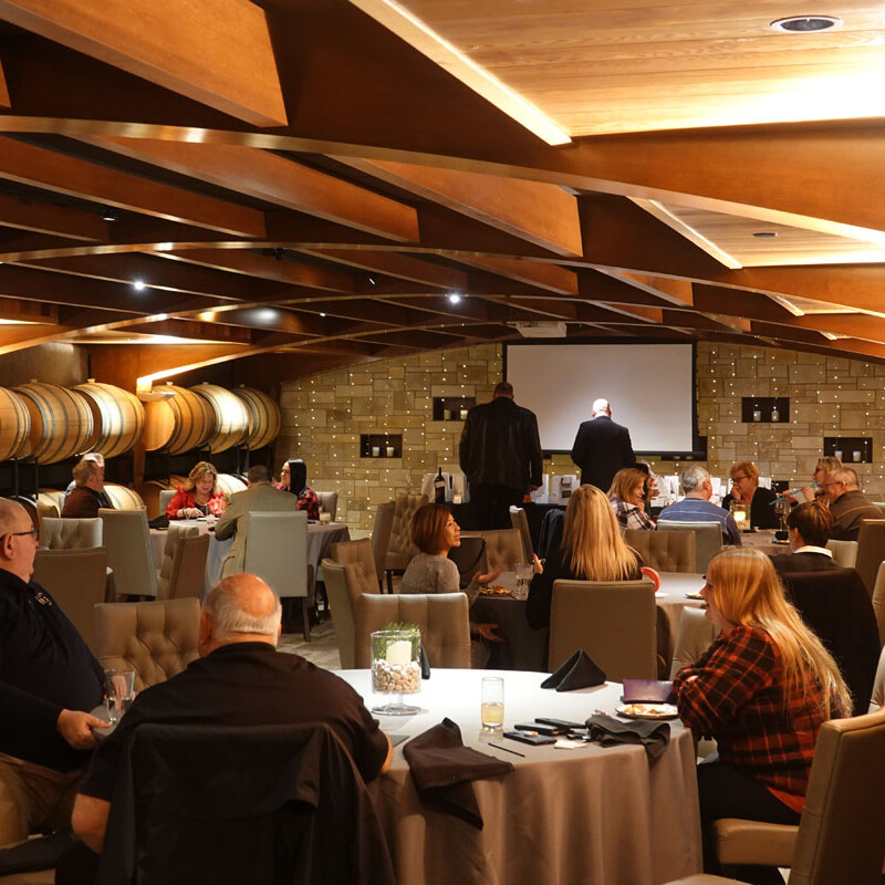 People are seated at round tables in a warmly lit room with wooden architecture and barrels along the walls. They appear to be engaged in conversation or listening to a speaker at the front, where a projector screen displays "Holiday Party 2023.