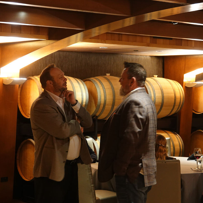 Two men in suits are engaged in a lively conversation in a room with wooden barrels stacked against the walls. Other people, possibly enjoying a holiday party, can be seen seated with wine glasses on tables in the background, creating an atmosphere that suggests a winery or wine cellar setting.