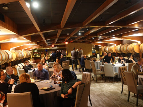 A lively restaurant filled with people seated at various tables, enjoying food and conversations, reminiscent of a festive holiday party. The ceiling has wooden beams, and large barrels line the back wall. The warm lighting creates a cozy ambiance perfect for memorable celebrations in 2023.