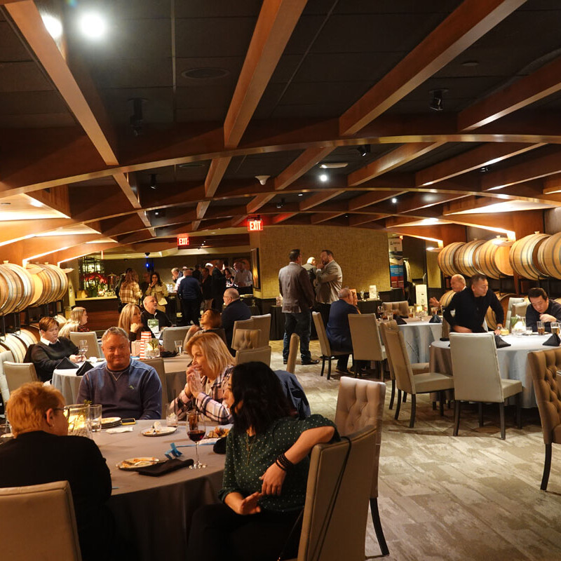 A lively restaurant filled with people seated at various tables, enjoying food and conversations, reminiscent of a festive holiday party. The ceiling has wooden beams, and large barrels line the back wall. The warm lighting creates a cozy ambiance perfect for memorable celebrations in 2023.