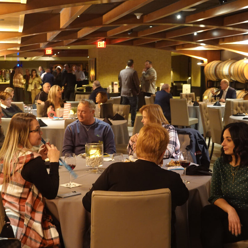 A group of people seated around a table engaging in conversation at a warmly lit restaurant with barrel décor, giving off a festive party vibe. Other patrons and servers can be seen in the background, contributing to the lively 2023 atmosphere. Glasses and plates are on the tables, suggesting a dining setting.