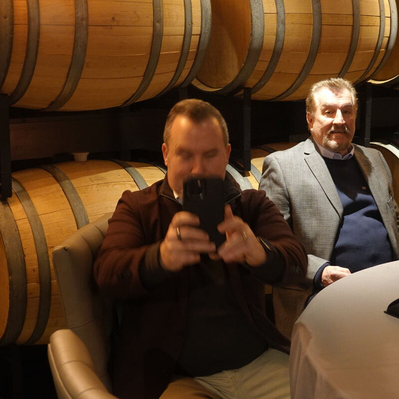 Two men are seated at a table in front of stacked wooden barrels. The man on the left, in a brown jacket, is holding up a phone, possibly taking a photo or video to capture the 2023 holiday moments. The man on the right, in a grey blazer, is looking at the camera. A drink is on the table.