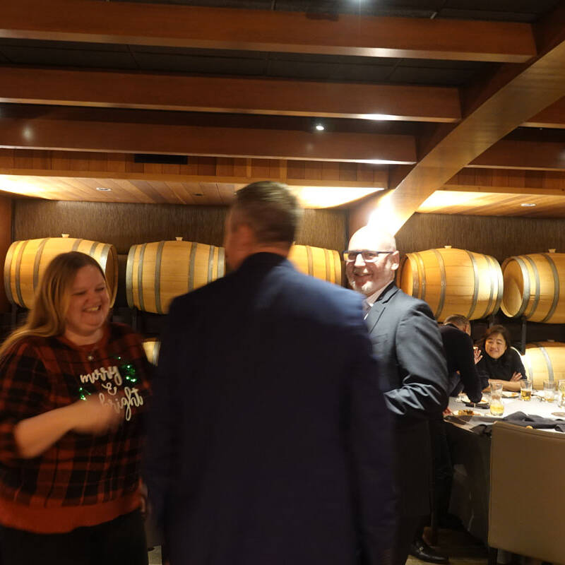 At a 2023 holiday party, a group of people are socializing in a warmly lit room with wooden barrels on the walls. A woman in a red plaid sweater and two men in blue and black suits are engaged in conversation. Other guests are seated at a round table in the background, enjoying drinks.