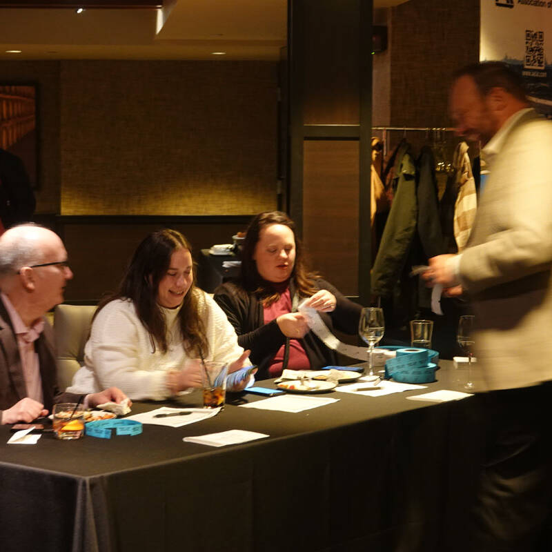 A group of people sit at a table handling raffle tickets inside a warmly lit room during the 2023 holiday party. One person is passing by the table, which has some drinks, a plate of food, and raffle items. A coat rack is visible in the background.