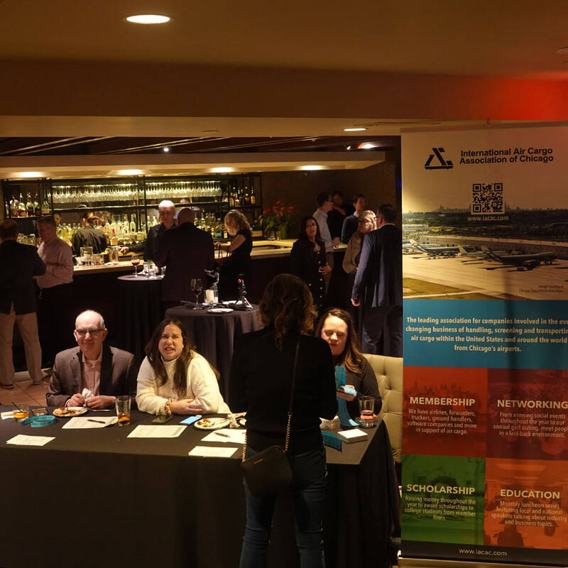 A group of people is gathered in a warmly lit conference room. In the foreground, three individuals sit at a table with informational materials. Behind them is a tall banner for the International Air Cargo Association of Chicago's 2023 holiday party. People mingle near a bar in the background.