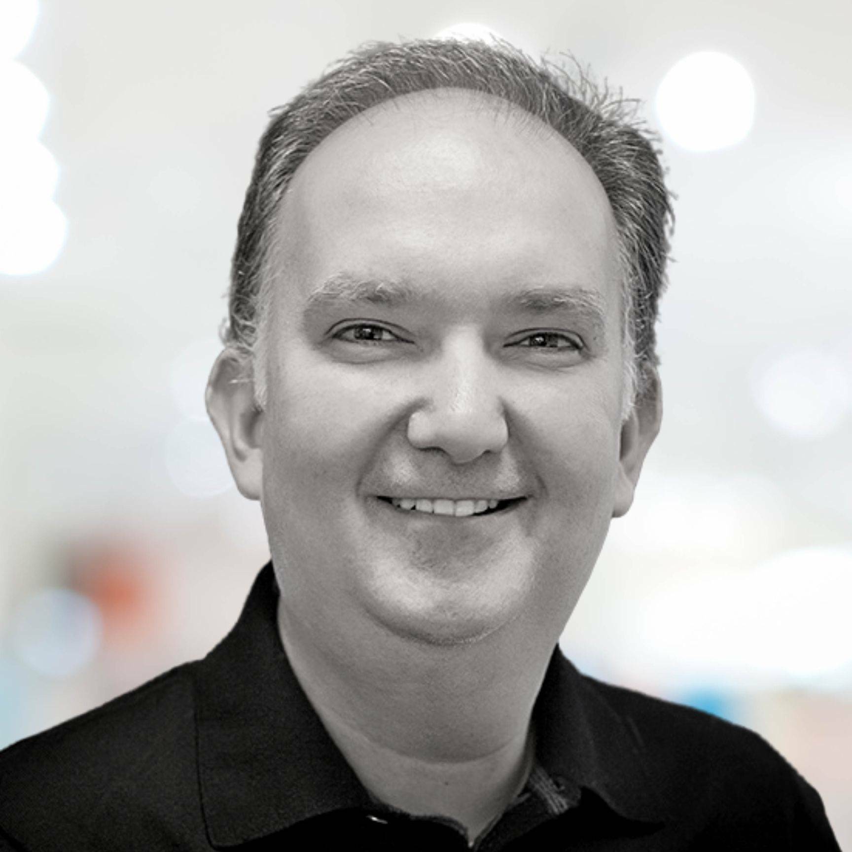 A smiling middle-aged man with short, dark hair wearing a black collared shirt. The background is softly blurred with a mix of light tones.