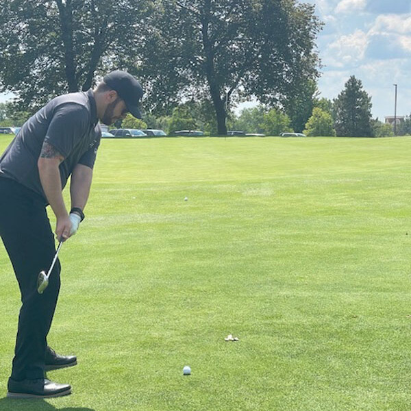 A man dressed in a dark shirt, pants, and cap is preparing to hit a golf ball on a grassy golf course during a sunny 2024 outing. Trees and parked cars are visible in the background under a partly cloudy sky.