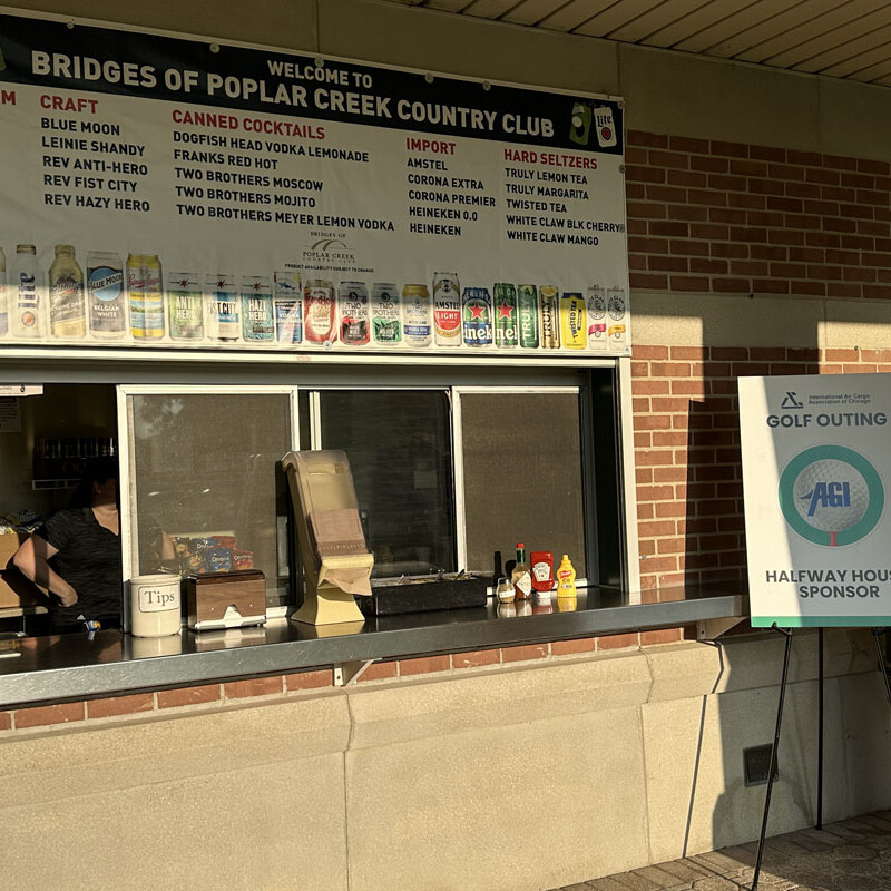 An outdoor concession stand with a large menu sign above offering various beers, cocktails, and drinks is located at the Bridges of Poplar Creek Country Club. Adjacent, a sign promotes an upcoming golf outing sponsored by AEC in 2023. The counter has condiments and a tip jar.