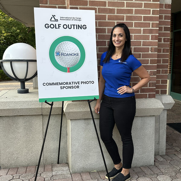 A woman in a blue polo shirt and black pants stands smiling next to a sign on an easel. The sign reads "2023 GOLF OUTING" and "COMMEMORATIVE PHOTO SPONSOR," with a circular logo that says "ROANOKE" in the center. The background includes a brick wall and a lamp post.