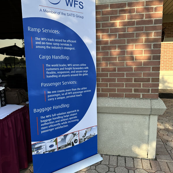 A vertical banner stands on a brick walkway, featuring the WFS logo at the top. It lists their services: Ramp Services, Cargo Handling, Passenger Services, and Baggage Handling, with brief descriptions underneath each heading. The background includes a building and a table with a tablecloth promoting their 2023 golf outing.