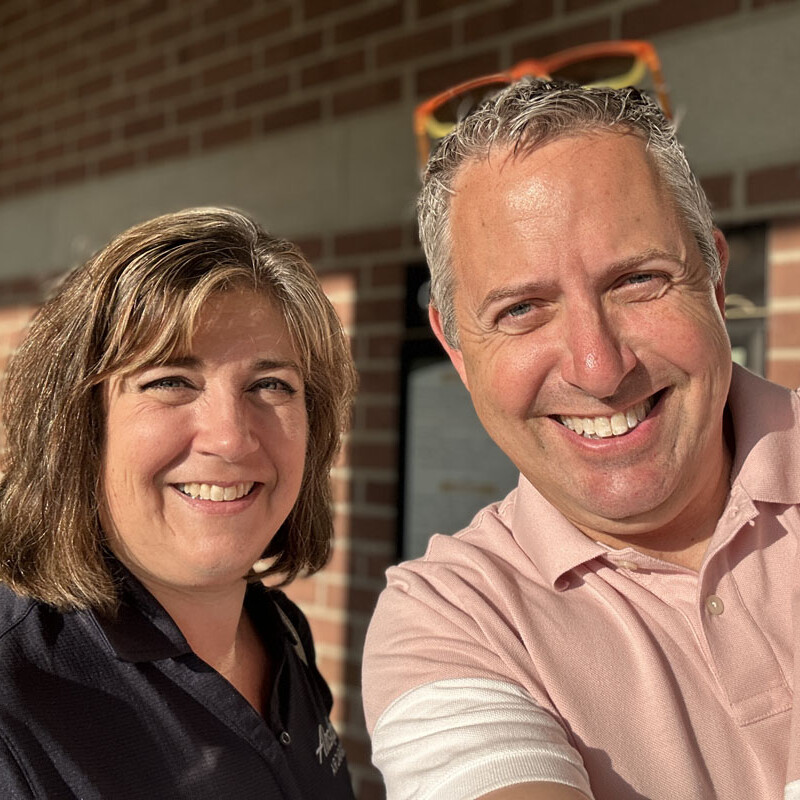 Two people are smiling and standing close together for a selfie during their outing in 2023. They appear to be outdoors in front of a brick building. The person on the left has short brown hair and is wearing a dark polo shirt, while the person on the right sports short gray hair and a pink polo shirt.