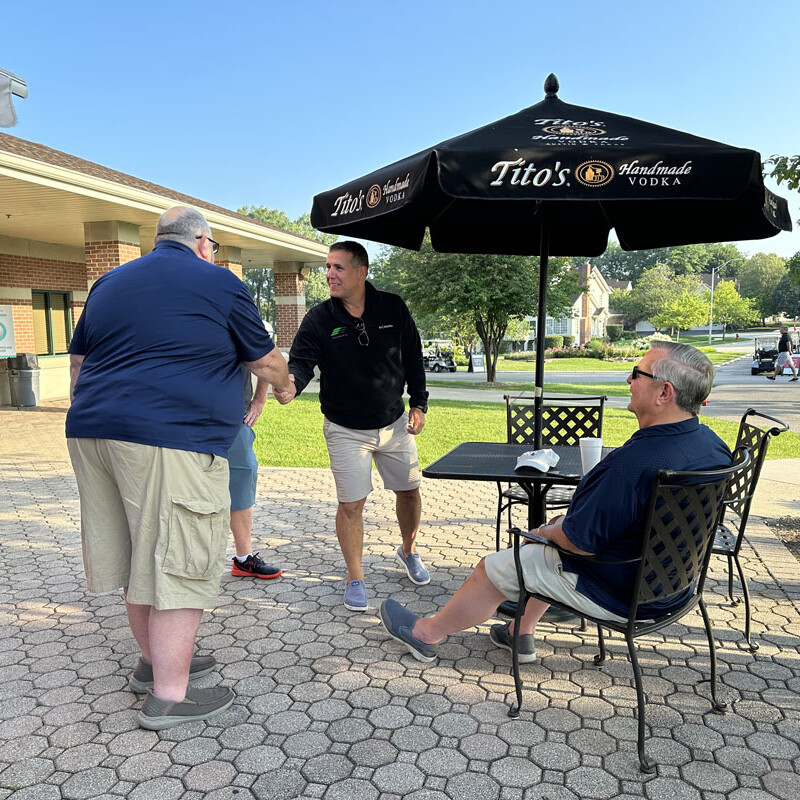 Four men are gathered outdoors near a shaded patio area with black metal furniture and a "Tito's Handmade Vodka" umbrella during the 2023 Golf Outing. Two men are shaking hands while another stands nearby, and a seated man in sunglasses observes the interaction. It is a sunny day.