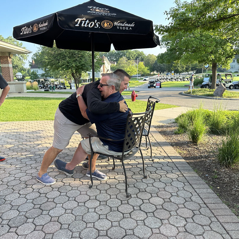 Two men are embracing under a tall black umbrella with the Tito's Vodka logo at a 2023 golf event. One man is seated while the other leans over from behind to give the hug. Another man stands nearby on a paved patio area surrounded by greenery and parked golf carts.