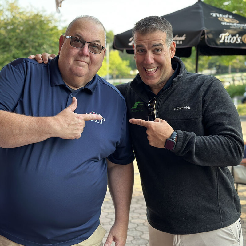 Two men are standing outside during a 2023 outing, both smiling and pointing at each other. The man on the left is wearing glasses and a navy shirt, while the man on the right has short hair and is dressed in a dark jacket. There is a man seated in the background under a black shade umbrella.