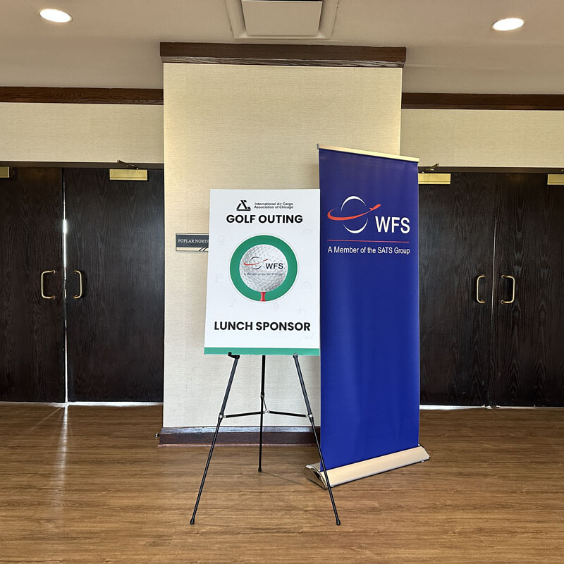 A stand with a "2023 Golf Outing" sign and "Lunch Sponsor" text is placed in front of double doors. The sign features a logo and details about the event. To its right is a blue banner with "WFS" and "A Member of the SFS Group" written on it. The wooden floor adds warmth to the setting.