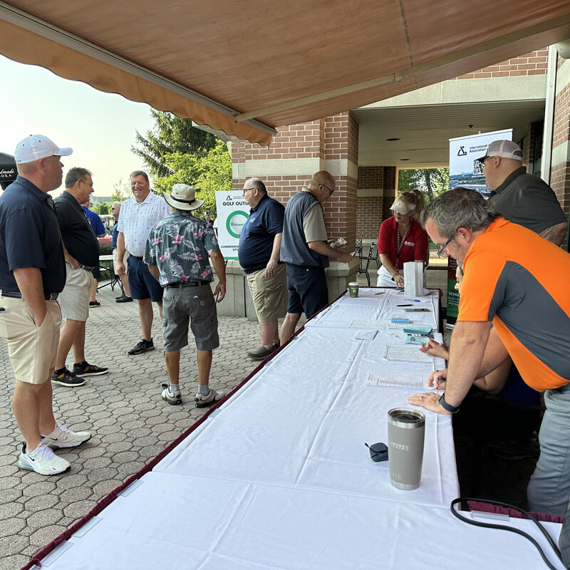 A group of people stand and interact at an outdoor registration table for the 2023 golf outing. The table is covered with a white cloth and various papers. Some are wearing golf attire and hats, contributing to the lively atmosphere. The setting is a brick building with a patio covered by an awning.