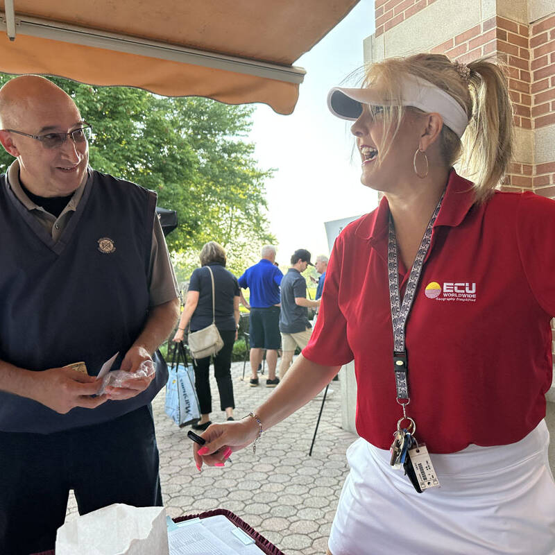 A woman in a red polo shirt and visor stands smiling with outstretched arms, talking to a bald man in a navy vest and glasses. They are at a 2023 golf outing under a canopy, with other people and a brick building visible in the background.