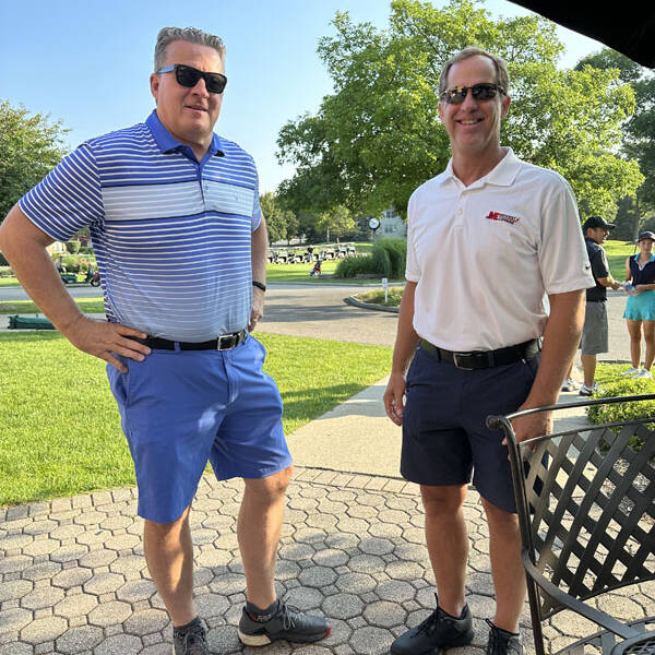 Two men in sunglasses, one in a blue striped polo and blue shorts, the other in a white polo and navy shorts, standing on a patio with brick pavers. Behind them are trees, grass, and other people enjoying a sunny day outing. A table with a chair is partially visible.