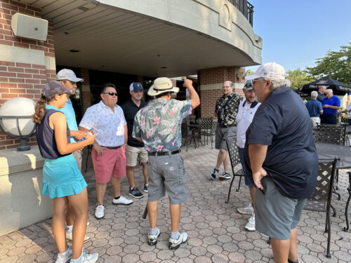 A group of eight people stand and chat outside a building with a covered patio during their 2023 outing. They are dressed in casual summer clothes, some in golf attire, and a couple of them wear hats. The weather appears sunny and clear, and there are tables and chairs on the patio.