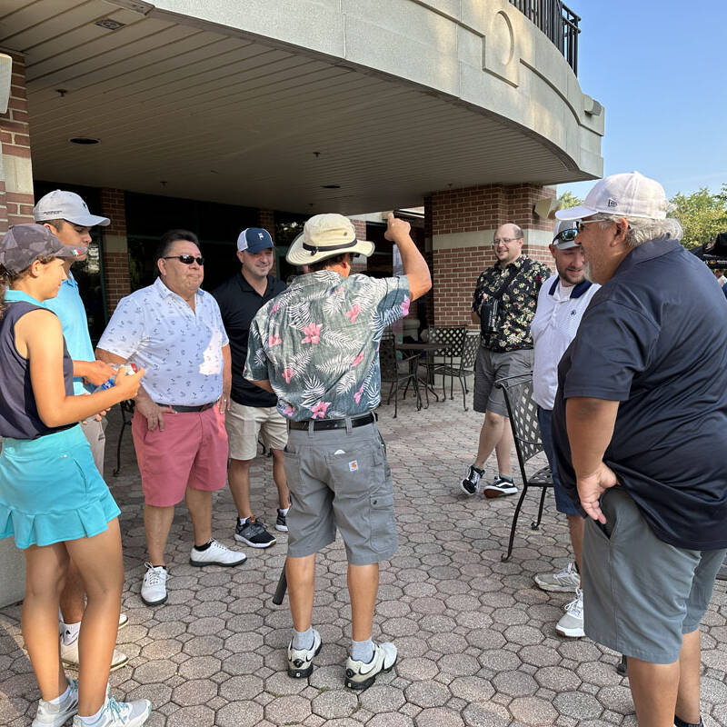 A group of eight people stand and chat outside a building with a covered patio during their 2023 outing. They are dressed in casual summer clothes, some in golf attire, and a couple of them wear hats. The weather appears sunny and clear, and there are tables and chairs on the patio.