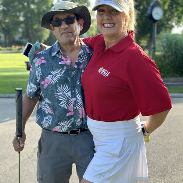 Two people are posing outdoors on a sunny 2023 day. The man on the left is wearing a tropical print shirt, sunglasses, and a hat, holding a golf club. The woman on the right is wearing a red polo shirt and white skirt with a visor, smiling with her arm around the man during their outing.